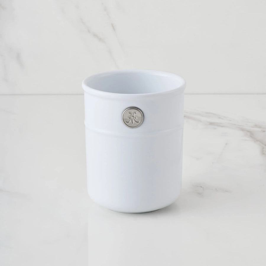 The Hestan Provisions Porcelain Bain Marie, featuring a silver circular emblem, rests on a white marble surface with a matching backdrop—an elegant touch for any kitchen.
