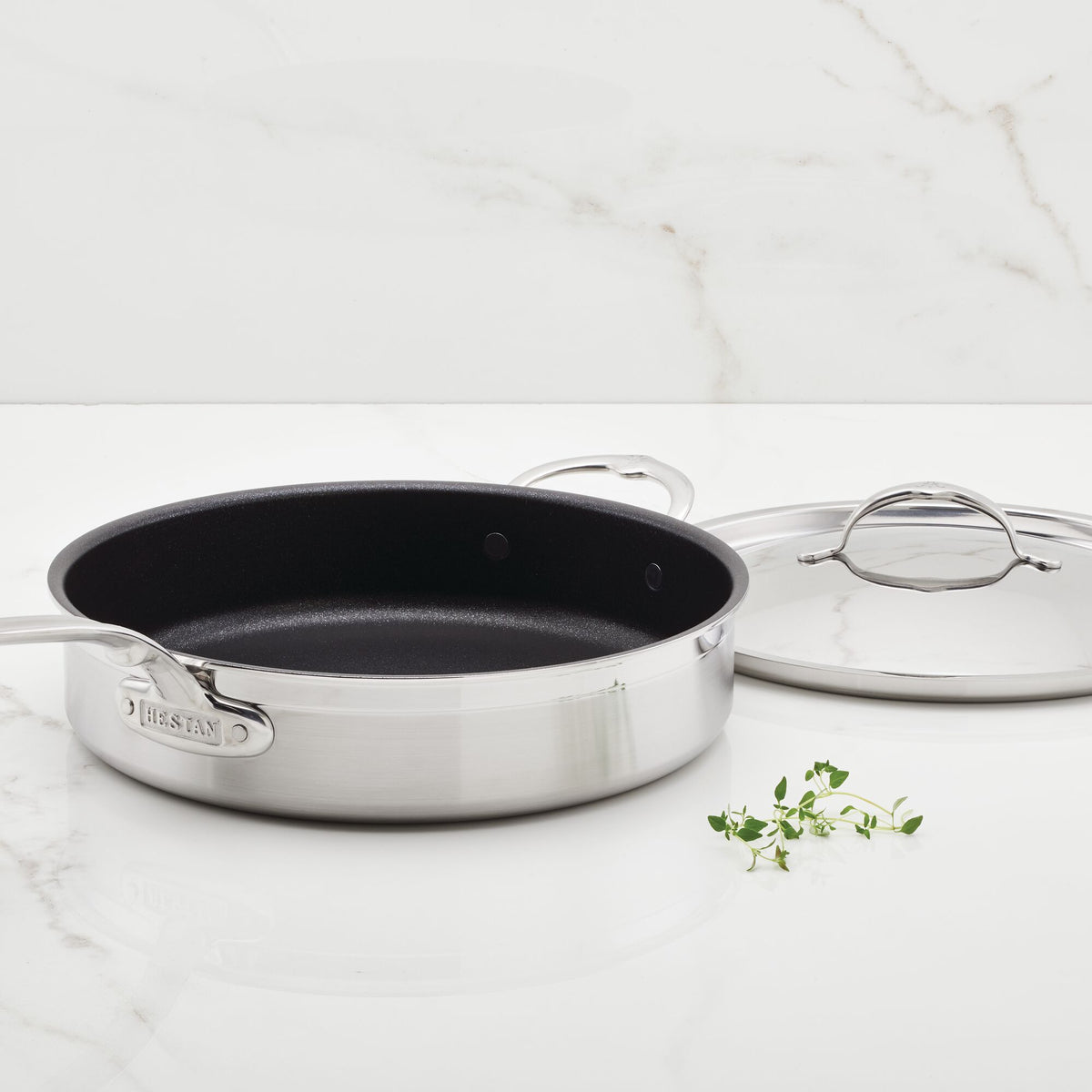 A ProBond Professional Clad Stainless Steel TITUM® Nonstick Sauté Pan with Lid, 5-Quart, sits on a marble countertop. The pan and its handle are paired with the lid beside it, while a small sprig of herbs enhances the sophisticated kitchen ambiance.