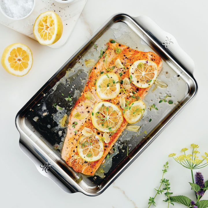 A baked salmon fillet topped with lemon, garlic, herbs, and black pepper is on a Hestan OvenBond Tri-Ply Quarter Sheet Pan & Rack (9x12 in), with halved lemons, salt dish, and fresh herbs on a white surface.