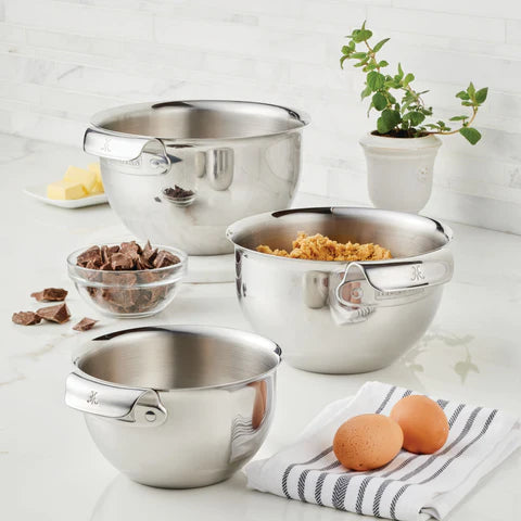 Three stainless steel mixing bowls on a countertop, containing spice, brown sugar, and a smaller bowl of chocolate chunks. Nearby are two eggs, a striped cloth, butter, and a potted plant. The background features a tiled wall.