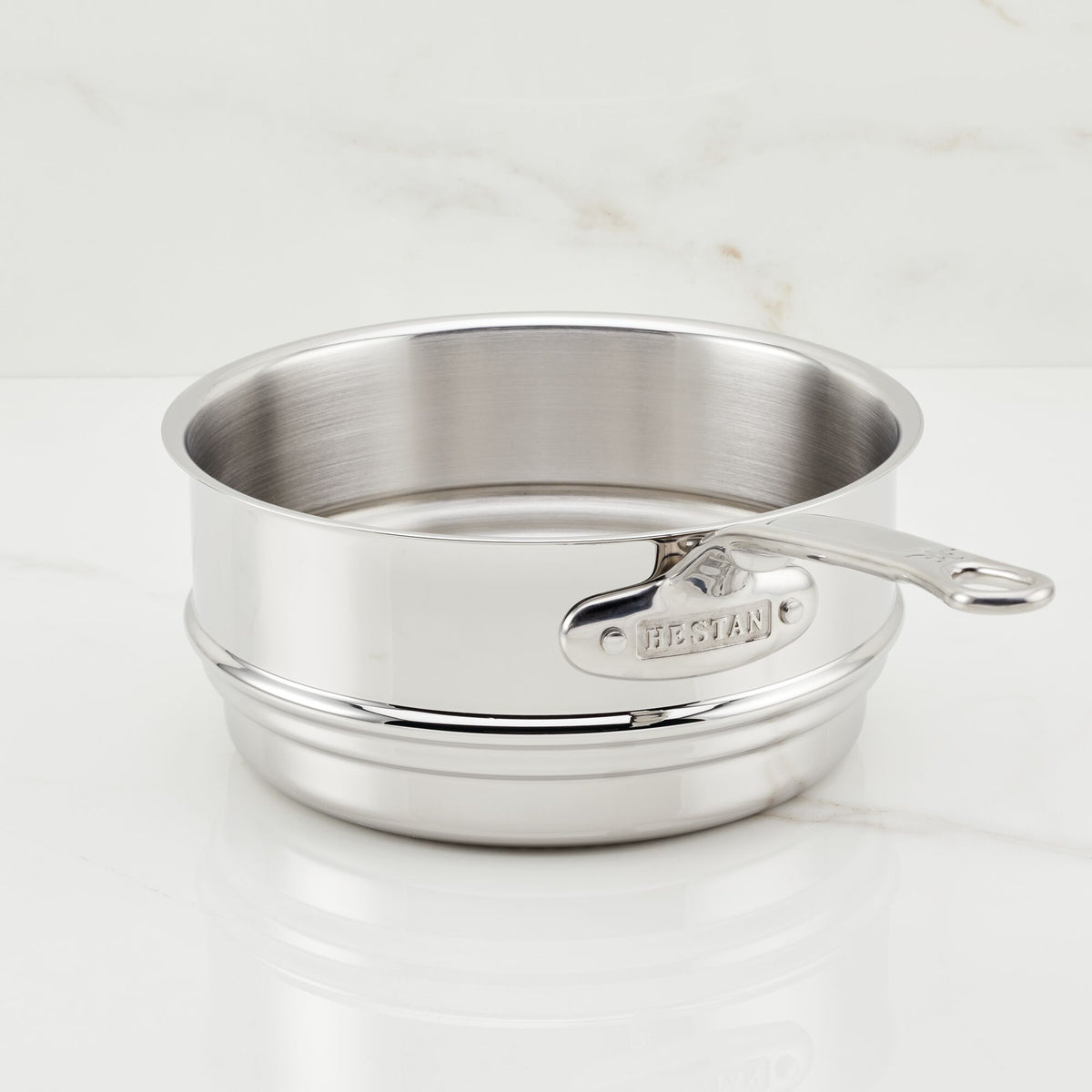 The Hestan Provisions Stainless Steel Steamer Insert, 3-Quart, with a handle, rests against a white marble background, displaying its raised brand logo on the side.