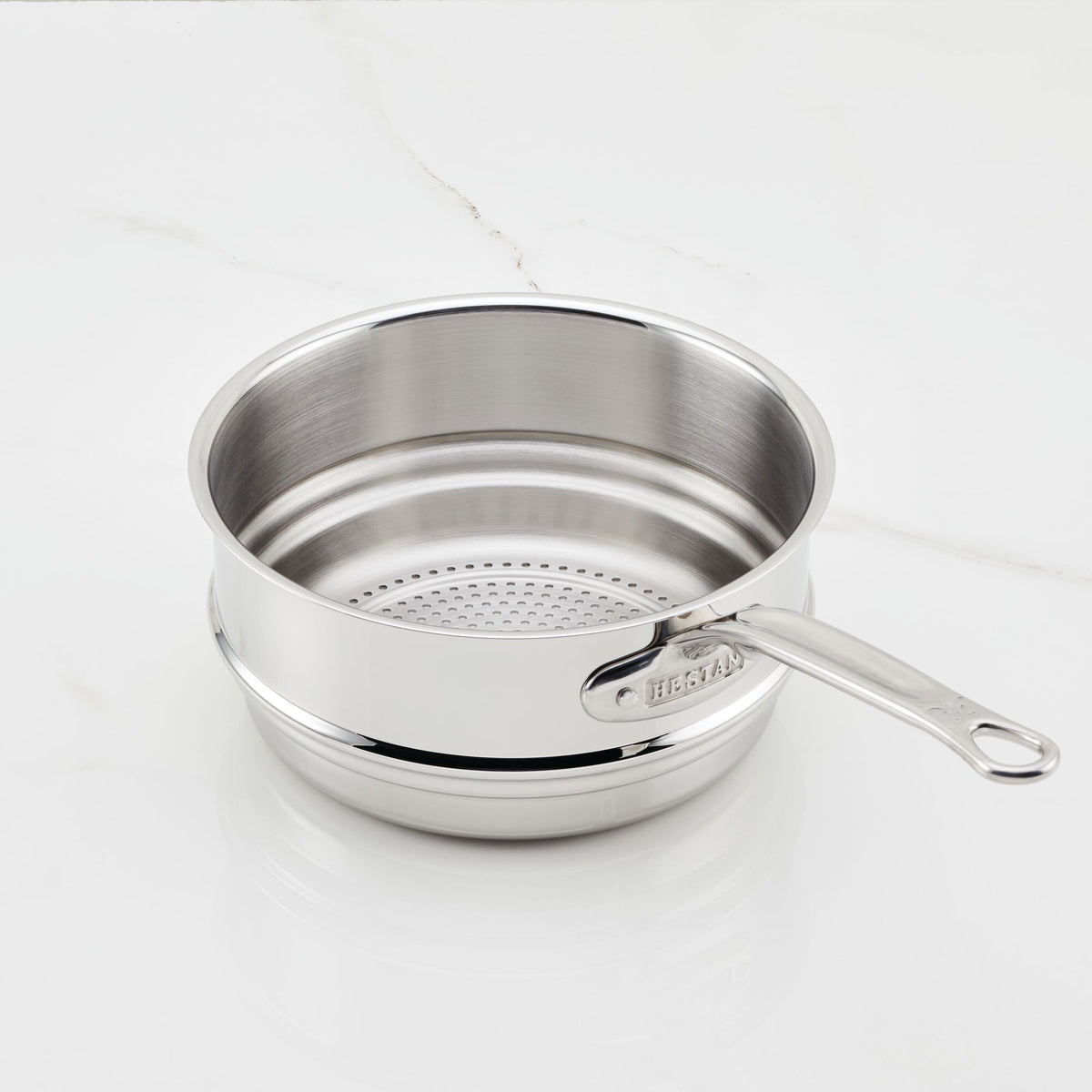 A Hestan Provisions Stainless Steel Steamer Insert, 3-Quart rests on a white marble surface and features a durable steaming insert with a perforated base. The long handle is elegantly branded.