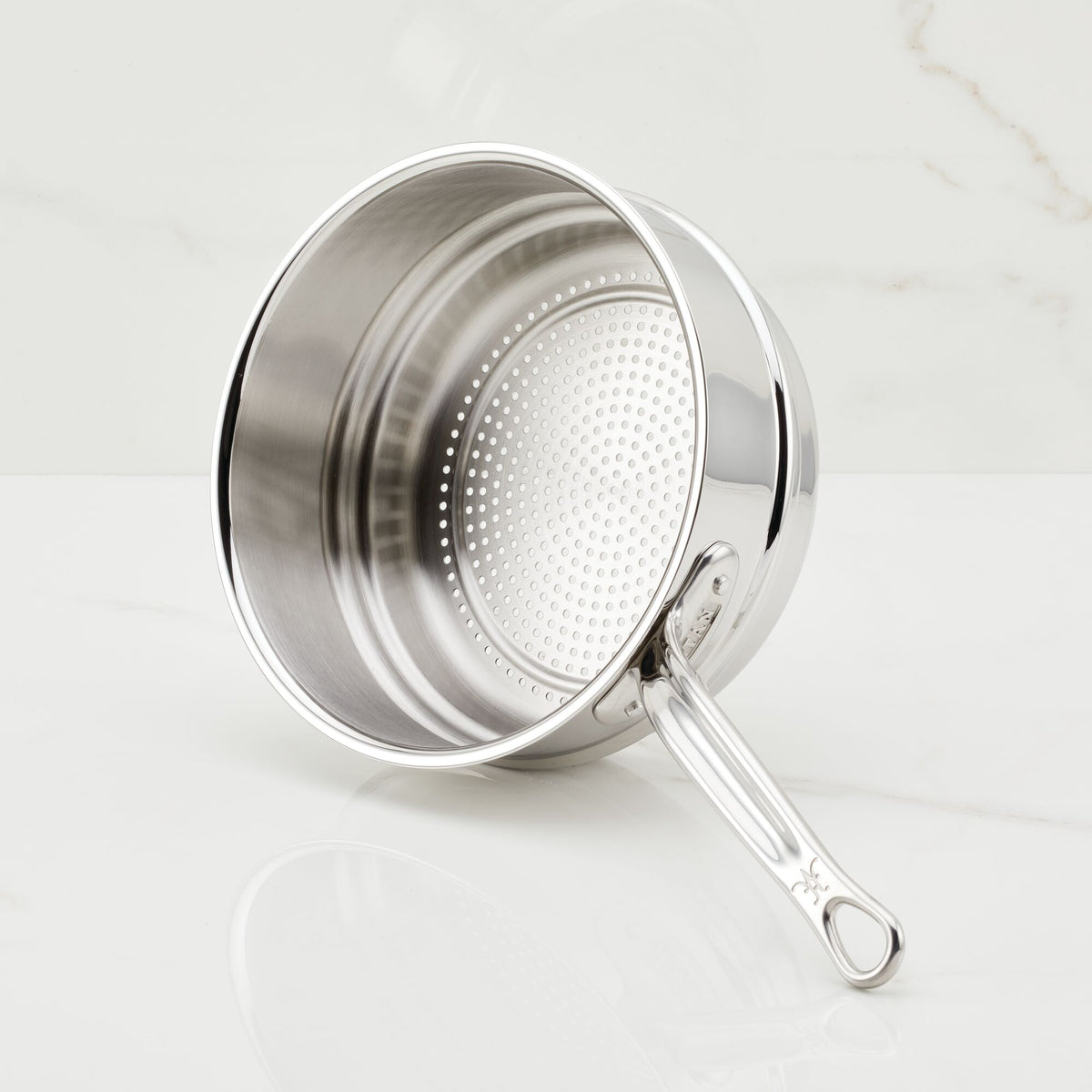 The Hestan Provisions Stainless Steel Steamer Insert, 3-Quart, sits on a white marble surface. It has a perforated base, rivet-mounted handle with a hanging loop, and a reflective interior/exterior for durability and style.