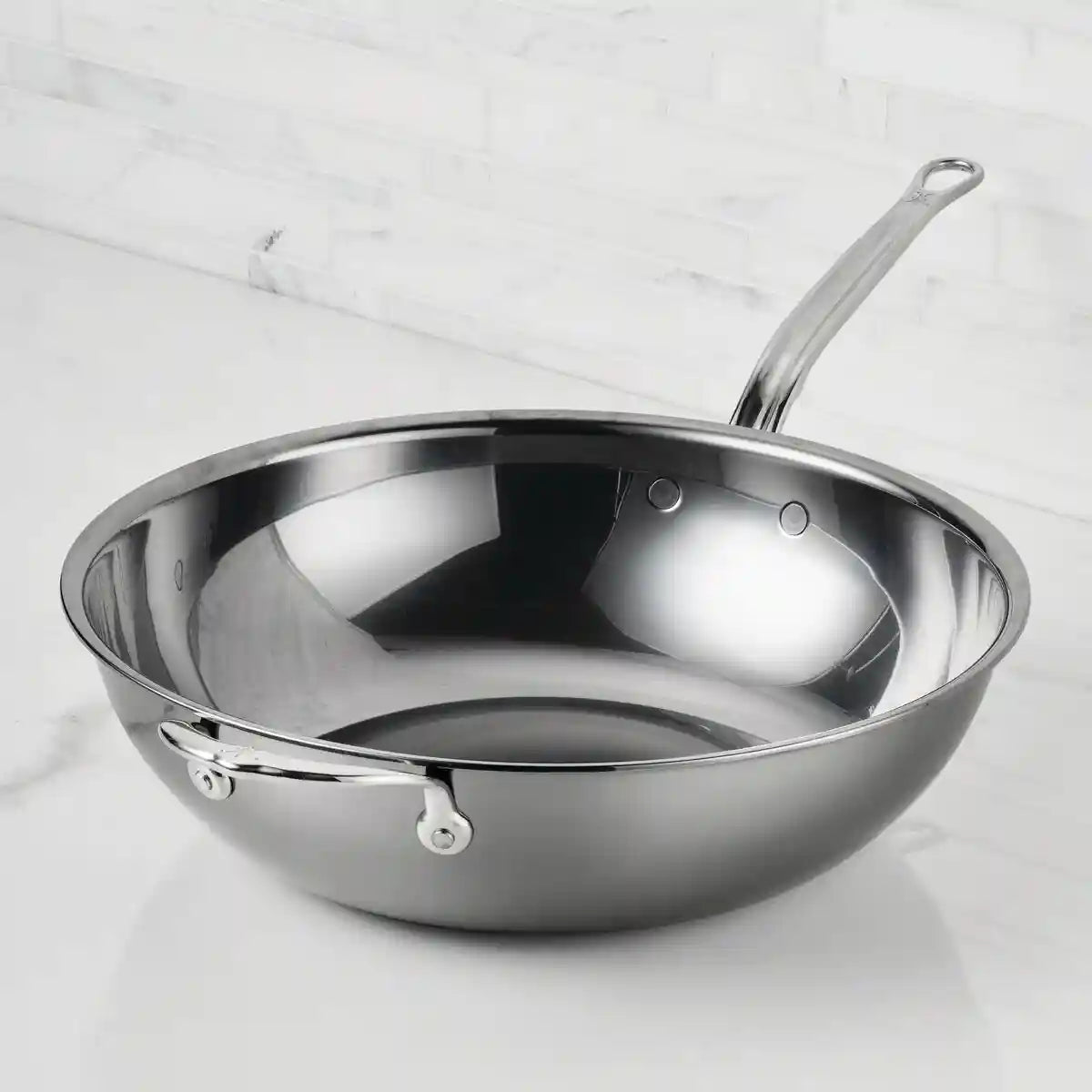 A NanoBond Titanium Chefs Pan, 14-Inch, with a shiny stainless steel finish and long and helper handles, sits on a white marble countertop against white tiles. Its molecular titanium construction ensures added durability.
