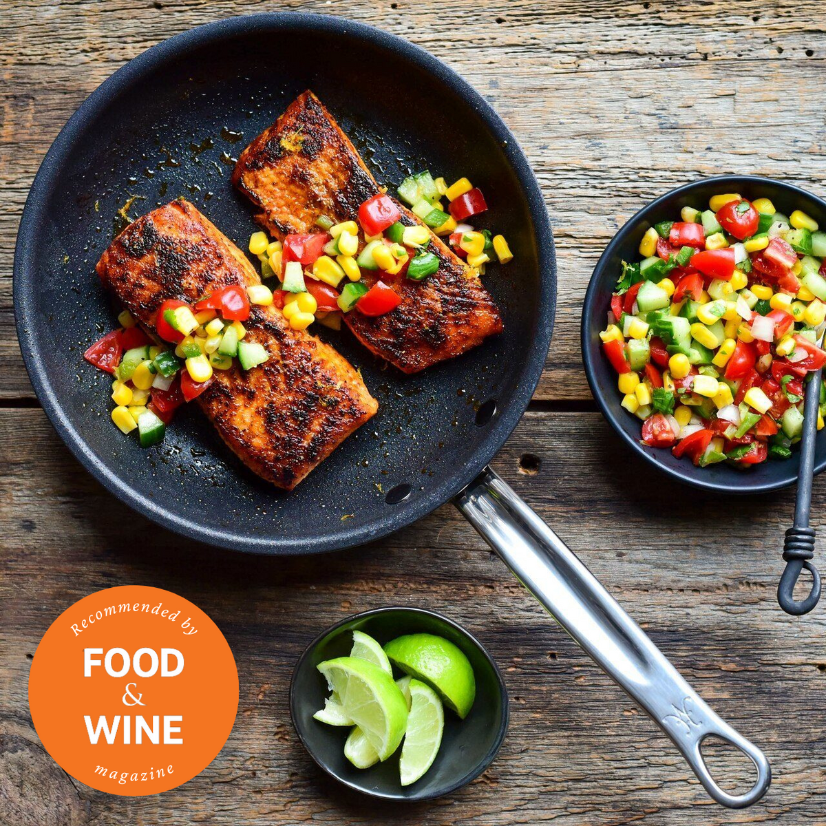 A ProBond Professional Clad Stainless Steel skillet from the 4pc Induction Cookware Set on a wooden table holds salmon with diced tomatoes, cucumbers, and yellow peppers. A dish of extra veggies and a lime wedge bowl sit nearby. A Recommended by FOOD & WINE magazine badge is visible.