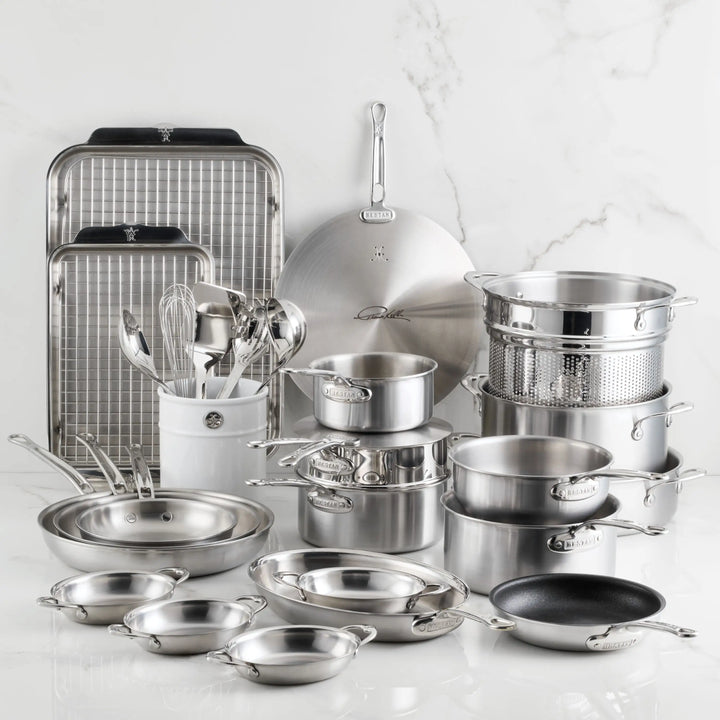 The Thomas Keller Insignia Commercial Clad Stainless Steel 29-Piece Cookware Set is displayed on white marble and features saucepans, frying pans, baking sheets, a steamer insert, plus metal whisks and spoons in a ceramic holder.