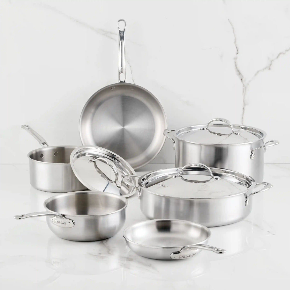 The Thomas Keller Commercial Clad Stainless Steel 9-Piece Cookware Set features saucepans, frying pans, and stockpots with lids, all arranged on a white surface against a marble background for versatile cooking.