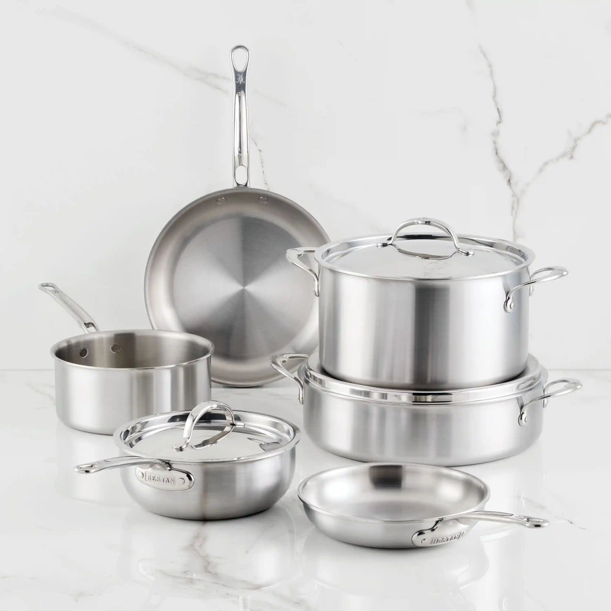 The Thomas Keller Commercial Clad Stainless Steel 9-Piece Cookware Set includes two saucepans, two frying pans, a sauté pan, and a stockpot with lids, elegantly displayed on a white marble countertop with a matching background.