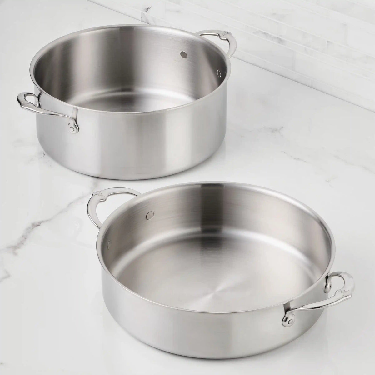 Two Thomas Keller Commercial Clad Stainless Steel Rondeaus, one deeper and one wider, with handles and a brushed metal finish, sit on a white marble countertop near a tiled backsplash.
