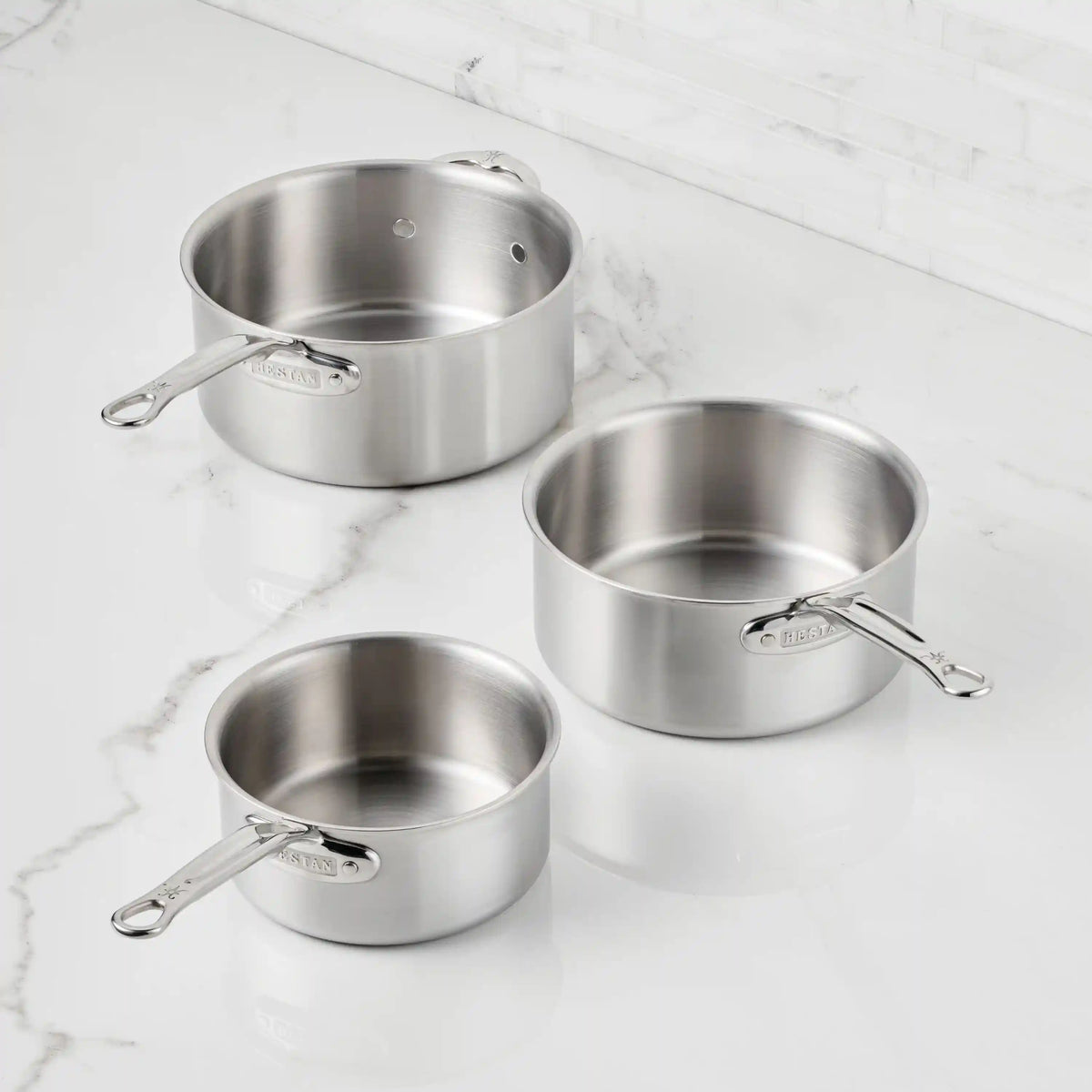 Three Thomas Keller Commercial Clad stainless steel saucepans from the 11-Piece Cookware Set, each with metal handles, are displayed on a white marble countertop against a backdrop of white tiled wall, emphasizing their sleek, professional style.
