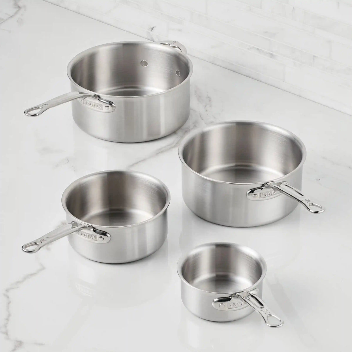 A Thomas Keller Commercial Clad Stainless Steel 0.75-Quart Butter Warmer with a metal handle sits empty on a white marble countertop, set against a white subway-tiled wall.