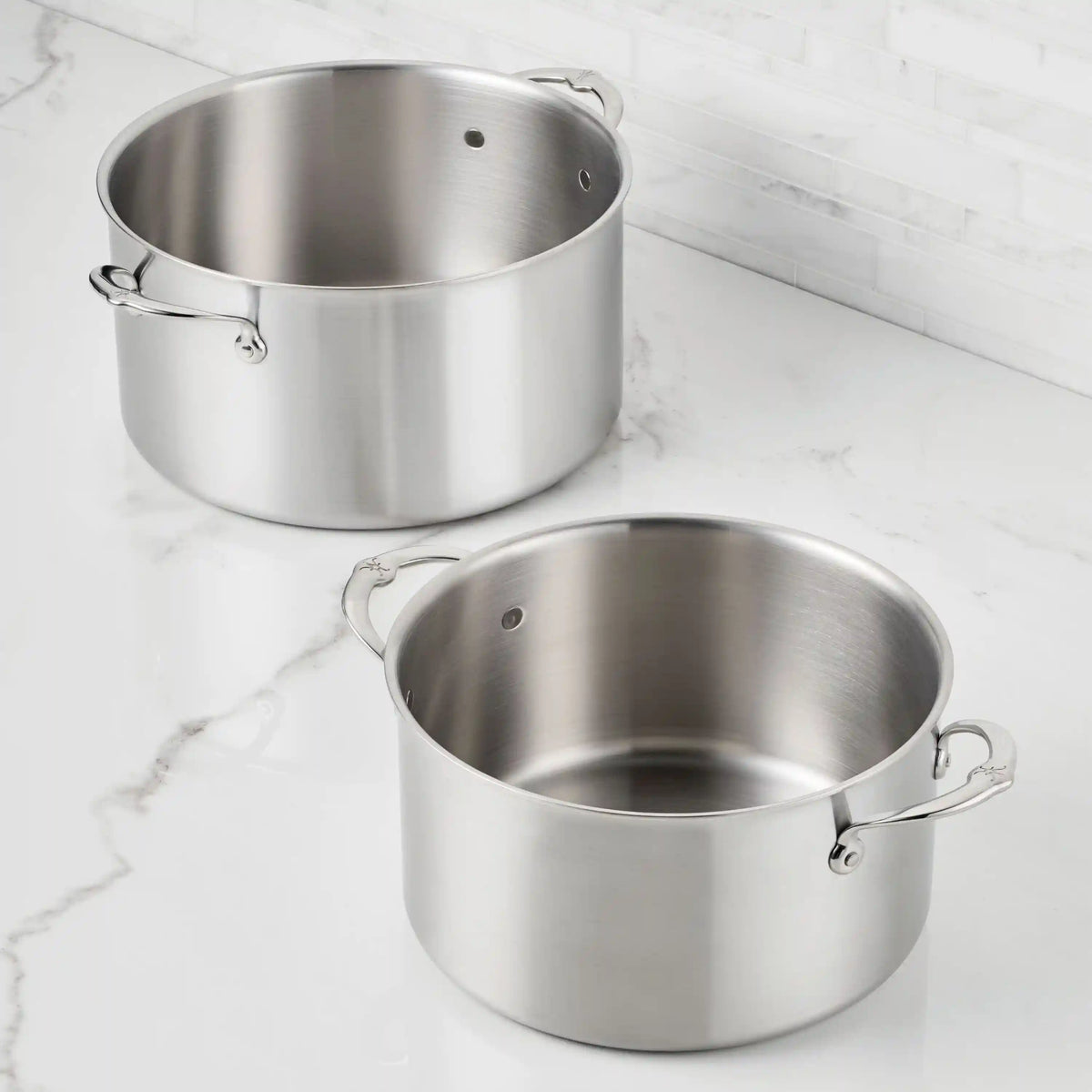 Two Thomas Keller Commercial Clad Stainless Steel Stock Pots with handles rest empty on a white marble countertop, their tri-ply construction ensuring professional culinary performance against a classic tiled backsplash.