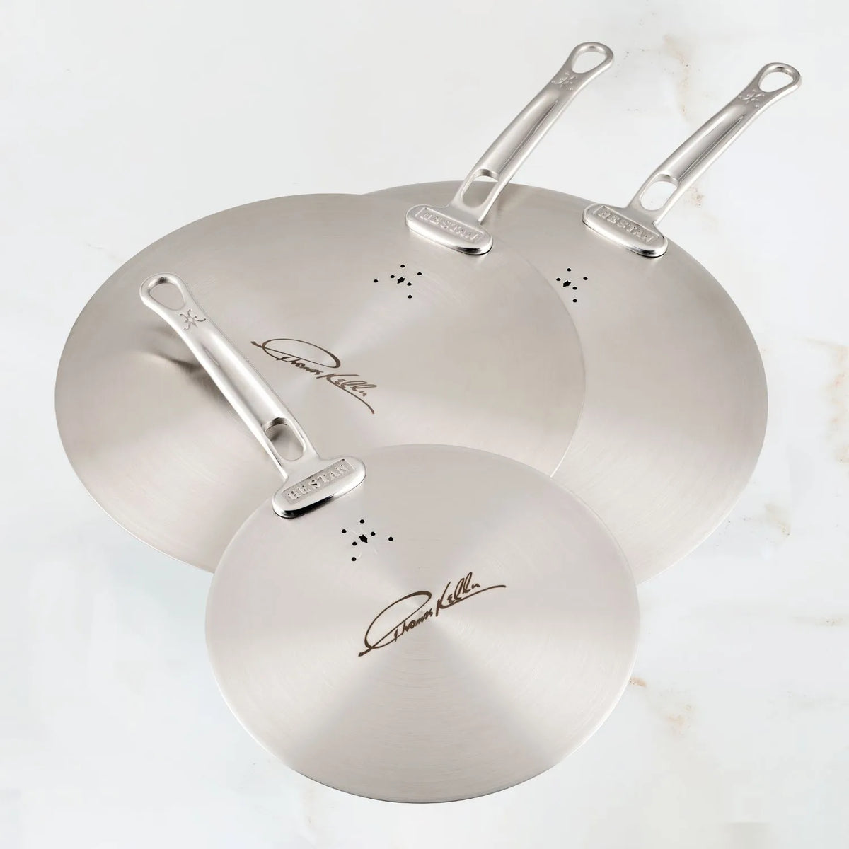 Three Thomas Keller Commercial Clad Stainless Steel Universal Lids with signature engravings, vent holes, and handles are arranged overlapping on a marbled surface—perfect for anyone seeking eco-friendly cookware.