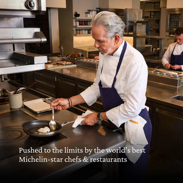 A chef in a white coat and navy apron cooks with the Thomas Keller Commercial Clad Stainless Steel 11-Piece Cookware Set in a pro kitchen. Another chef works behind. Text: Trusted by top Michelin-star chefs & restaurants worldwide.