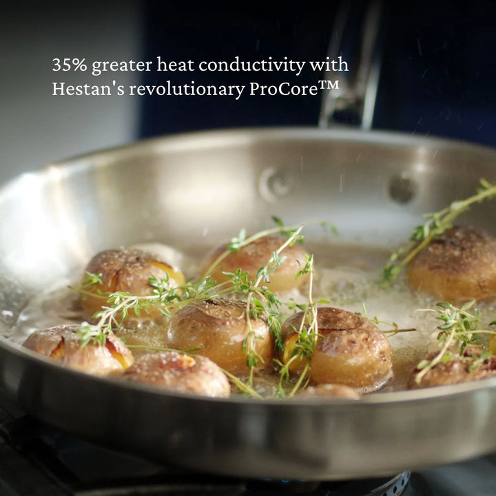 A tri-ply Thomas Keller Commercial Clad Stainless Steel 9-Piece Cookware Set frying pan cooks halved potatoes with herbs on the stove, overlaid with “35% greater heat conductivity with Hestan’s revolutionary ProCore™.”.