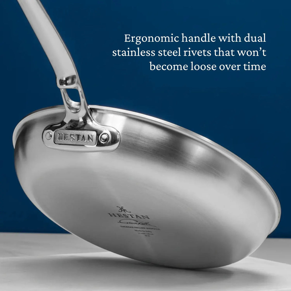 A close-up of the Thomas Keller Commercial Clad Stainless Steel 9-Piece Cookware Set showcases an ergonomic handle with dual rivets, emphasizing its tri-ply clad design for lasting durability and professional-grade performance against a blue background.