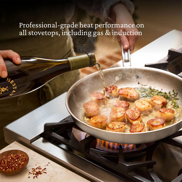 A person pours liquid into a stainless steel pan of seared scallops on a gas stove. Text: Achieve professional results with the Thomas Keller Commercial Clad Stainless Steel 9-Piece Cookware Set for all stovetops, including gas & induction.