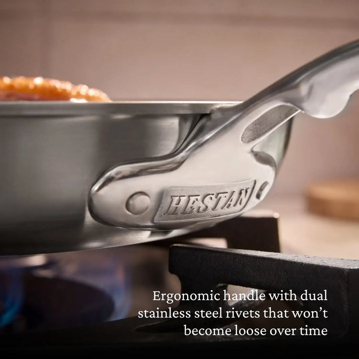 A close-up of a Thomas Keller Commercial Clad Stainless Steel 7-Piece Cookware Set pan with ergonomic, riveted handle on a stovetop burner. The handle’s base is stamped. Text: Professional performance for commercial kitchens.