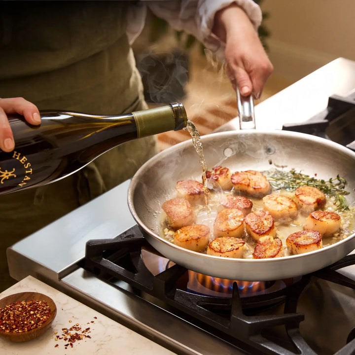 Someone pours white wine from a bottle into a hot skillet from the Thomas Keller Commercial Clad Stainless Steel 10-Piece Cookware Set with seared scallops and herbs on a gas stove, steam rising as a bowl of red pepper flakes sits nearby.