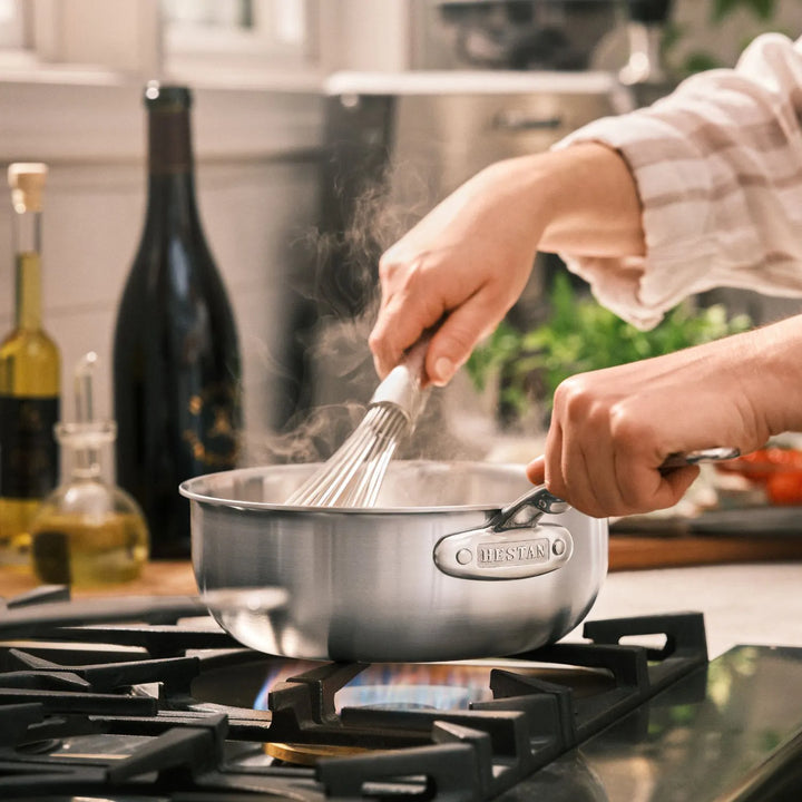 A person whisks food in a Thomas Keller Commercial Clad Stainless Steel pan from the 10-Piece Cookware Set on a gas stove, steam rising, with bottles of oil, wine, and fresh herbs visible in the background.