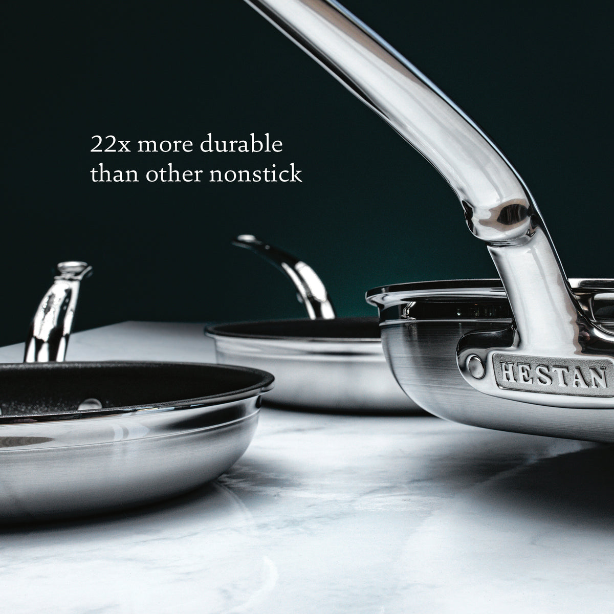 Close-up of three ProBond Professional Clad stainless steel frying pans on a marble surface. One pans handle, engraved with HESTAN, is in focus. The background features dark green with text claiming the set is 22x more durable than other TITUM® Nonstick.