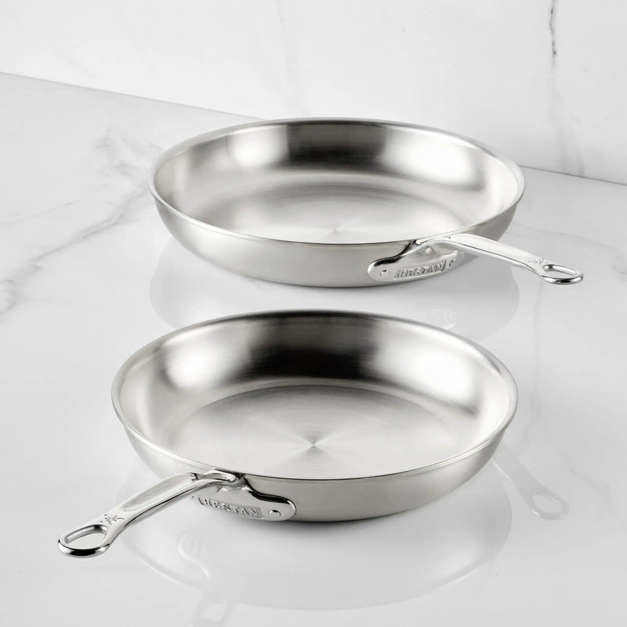 Two Thomas Keller Commercial Clad Stainless Steel Skillets with polished metal handles rest on a white marble surface, positioned at slight angles to showcase their tri-ply professional-grade design.