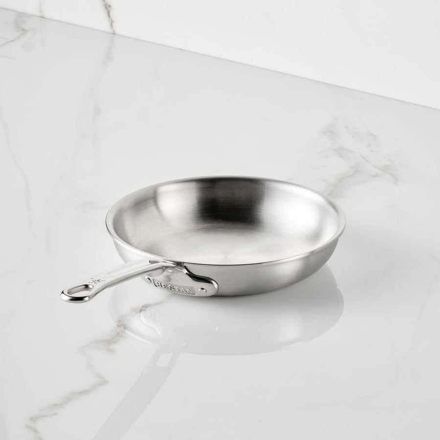 The Thomas Keller Commercial Clad Stainless Steel Skillets feature a chef-designed, long-handled tri-ply construction, shown angled on a white marble countertop with gray veining for a sleek kitchen aesthetic.