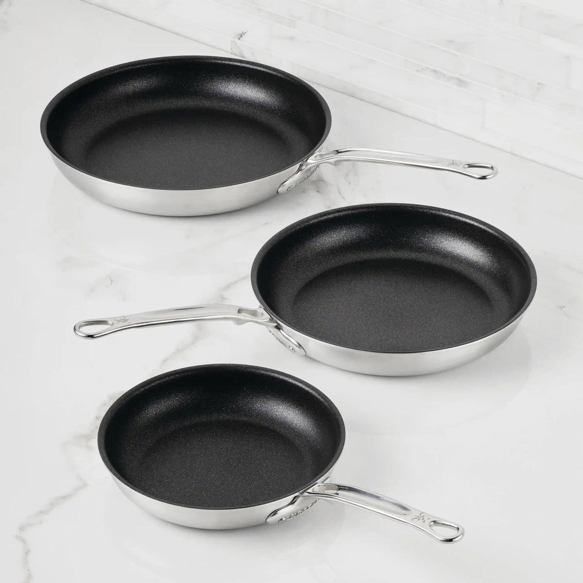 Three Thomas Keller Commercial Clad Stainless Steel TITUM® Nonstick Skillets are arranged by size on a white marble countertop, offering professional performance with their durable construction and sleek black interiors.