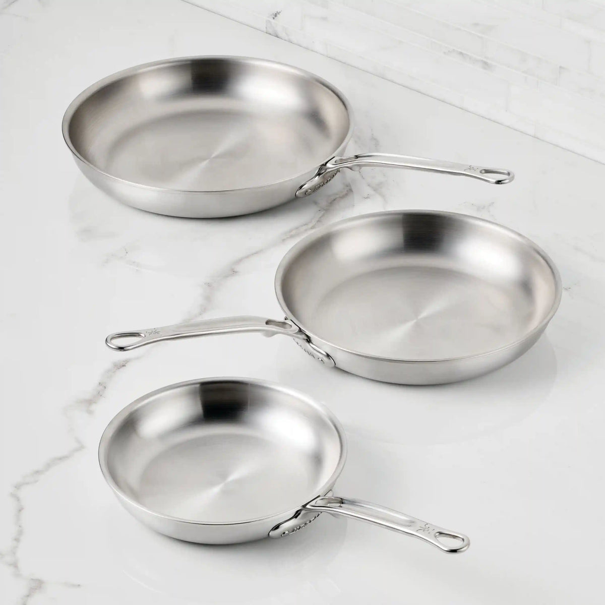 Three Thomas Keller Commercial Clad Stainless Steel Skillets in varying sizes, each with long handles and a brushed finish, are displayed on a white marble countertop with white tiled walls in the background.