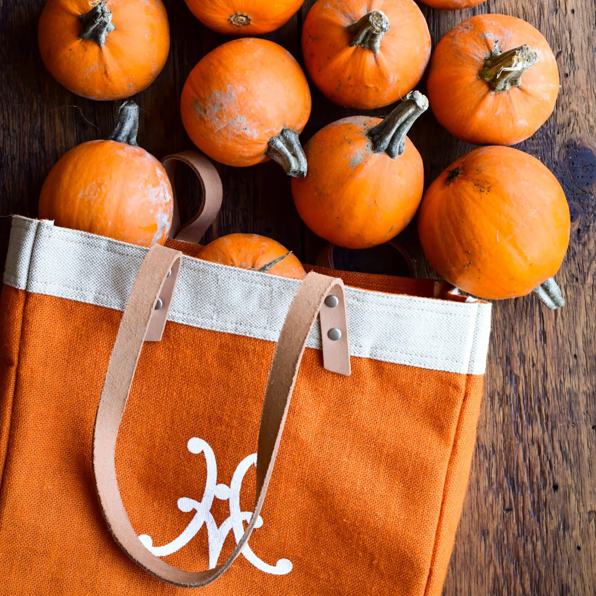 Several small orange pumpkins sit in and around the Accessories Hestan Market Tote in Citra Orange, which features beige leather shoulder straps and a white decorative “M” on the front, resting on a wooden surface.