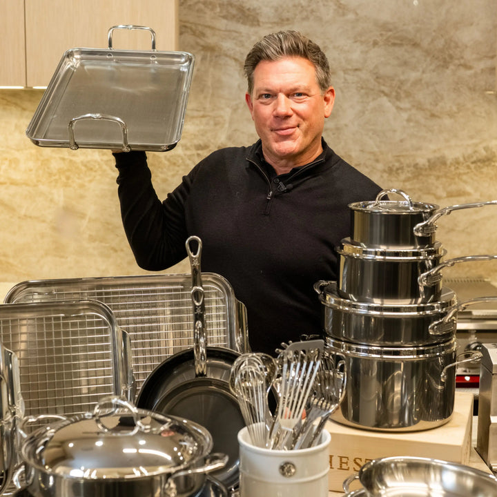 A man in a black shirt stands in a kitchen holding a metal tray. In front of him is the NanoBond Titanium x Tyler Florence Cookware Set, 41-Piece, with pots, pans, trays, and utensils neatly arranged on the counter.