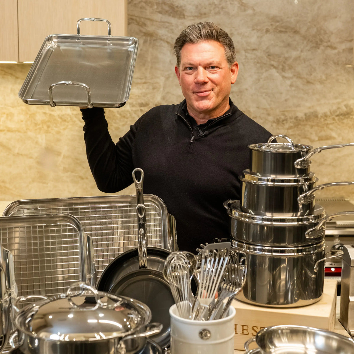 A man in a black shirt stands in a kitchen, holding up a metal baking tray. In front of him are various stainless steel pots, pans, lids, and whisks from the NanoBond® Titanium x Tyler Florence Cookware Set, 41-Piece—each piece neatly organized on the counter and celebrated for their distinctive Molecular Titanium finish.