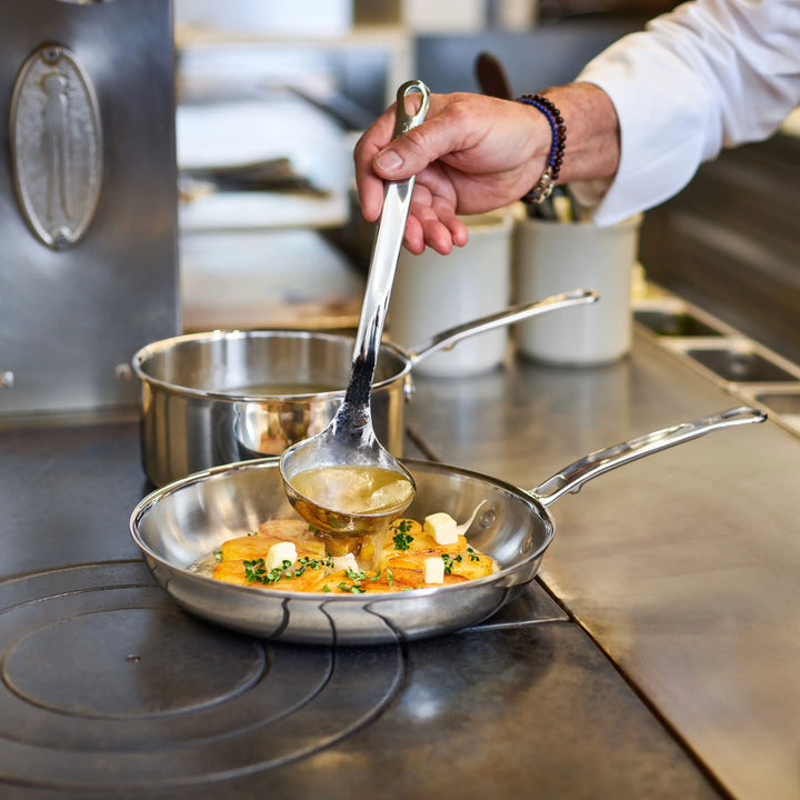 A person pours sauce from a ladle onto food cooking in ProBond® Luxe™ Polished Clad Stainless Steel Signature Set, 7-Piece cookware on the stovetop, with another pot and utensils visible in the background.