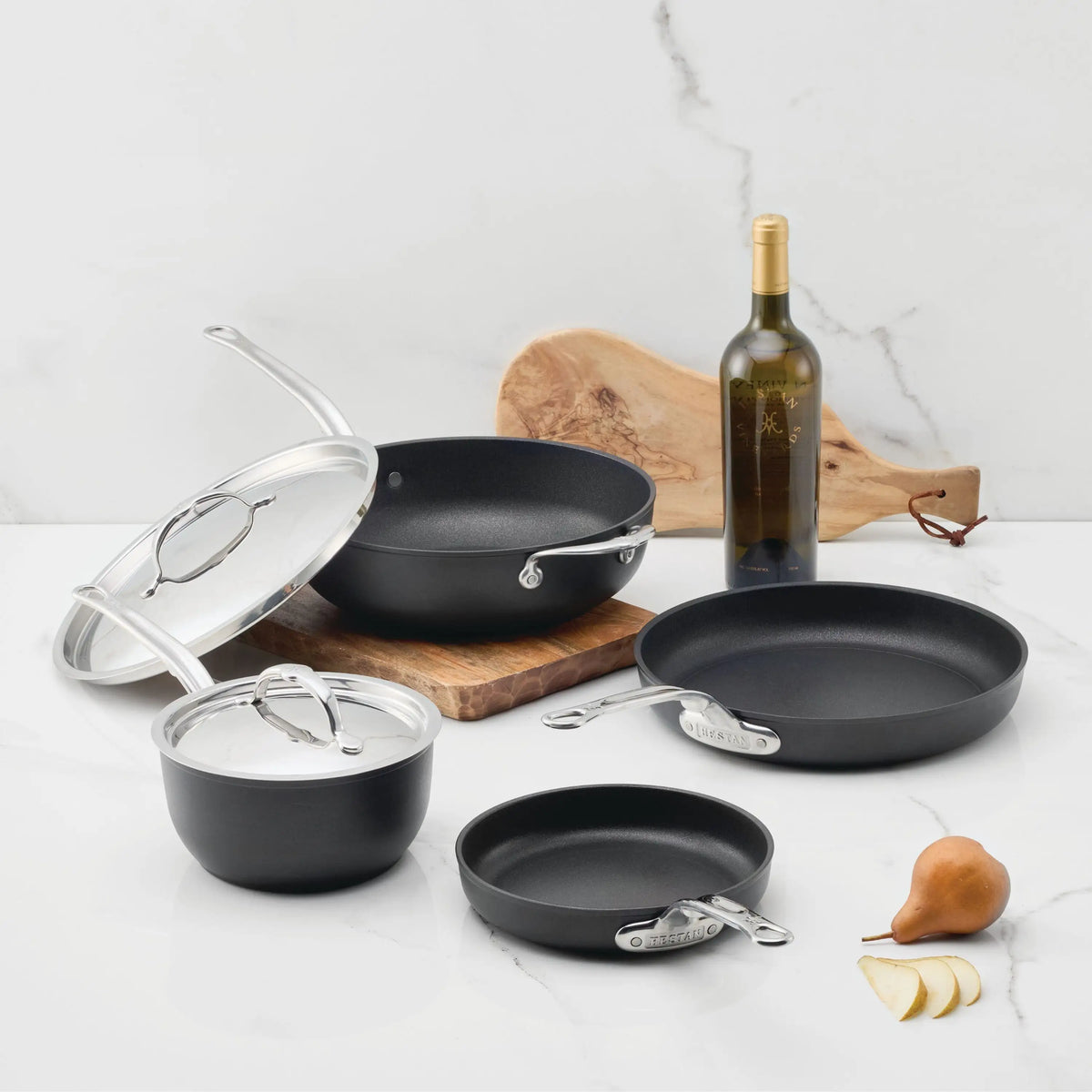 Four black TITUM® Fusion TITUM Nonstick Cookware Set, 6-Piece pieces with metal handles and lids are arranged on a white countertop, with a wooden cutting board, white wine bottle, and pears in the background.