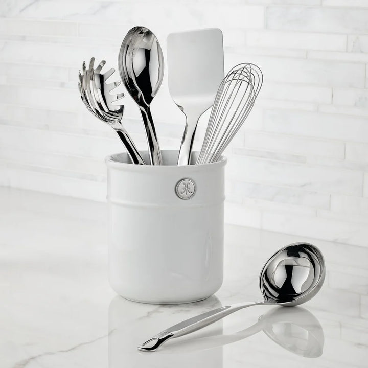 A Hestan Provisions Porcelain Bain Marie sits on a marble countertop holding five chef’s tools: spaghetti server, serving spoon, slotted spoon, spatula, and whisk. A ladle rests neatly on the counter beside the holder.