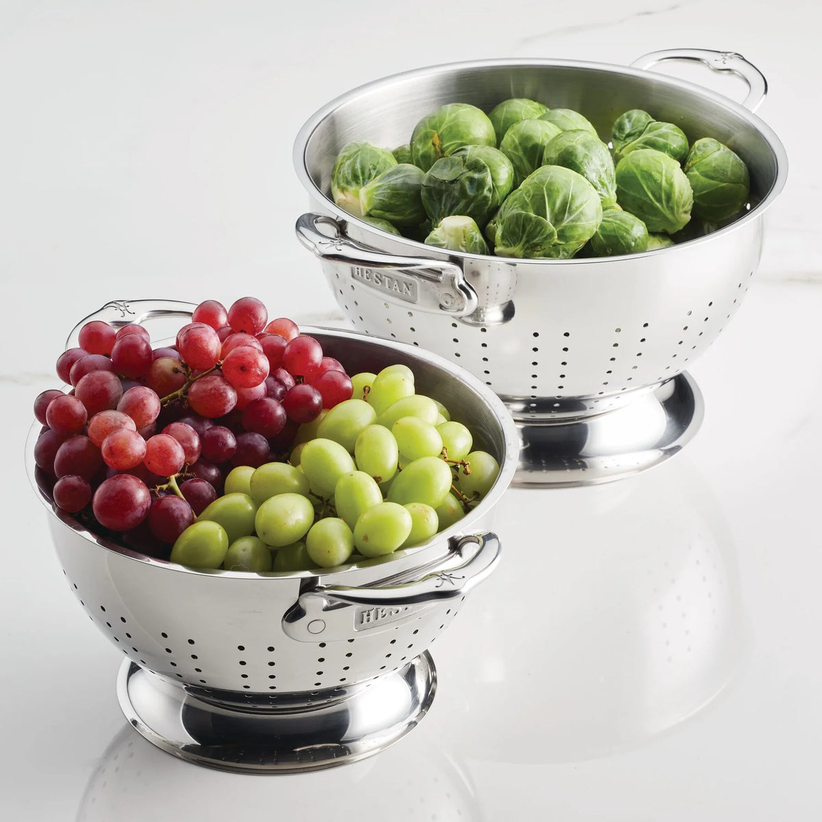 Two stainless steel colanders from the Hestan Provisions Host and Roast 8-piece set sit on a white surface—one with green and red grapes, the other with Brussels sprouts. The brand name is embossed on their handles.