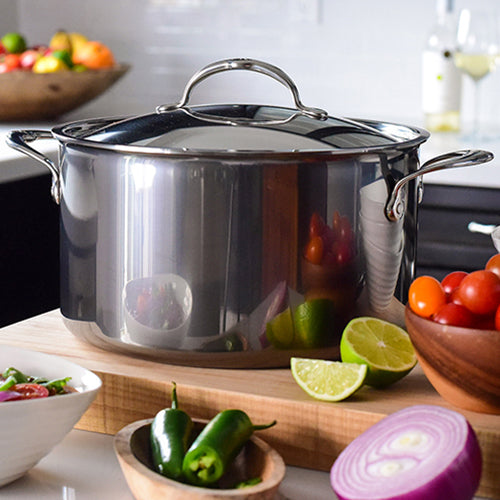 A sleek NanoBond Titanium Stockpot, 8-Quart with a lid sits on a wooden countertop, surrounded by ingredients like tomatoes, lime halves, jalapeños, and a red onion. In the background, more tomatoes rest in a bowl alongside a glass of white wine.