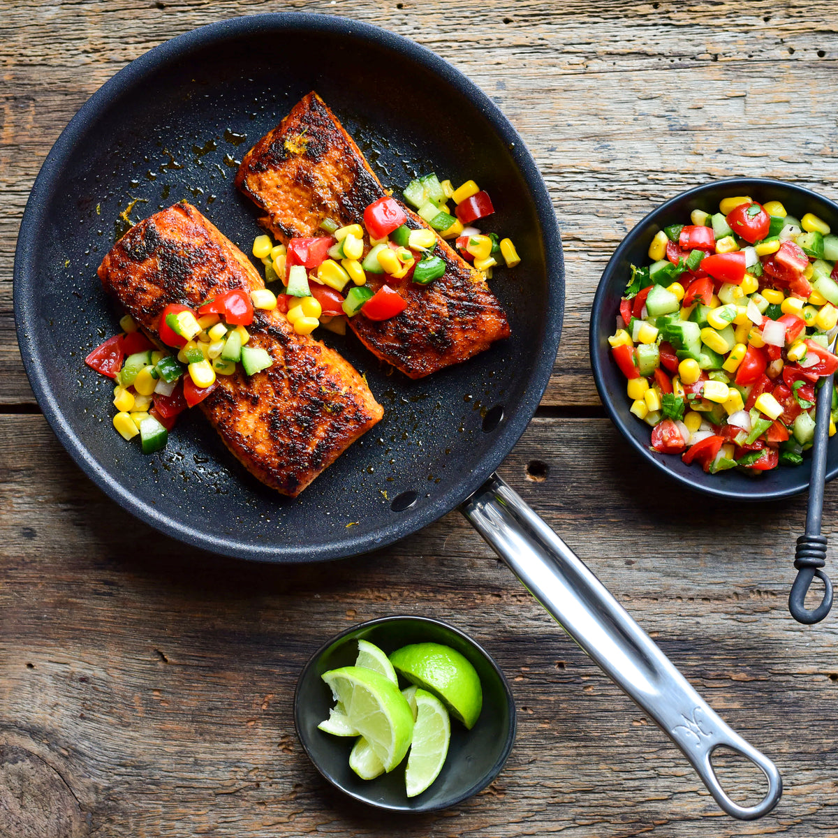A pan from the ProBond® Forged™ Professional Clad Stainless Steel Epicurean Set, 15-piece holds grilled salmon with tomato, cucumber, and corn salsa. Two bowls—one with extra salsa and one with lime wedges—sit nearby on the wooden surface.