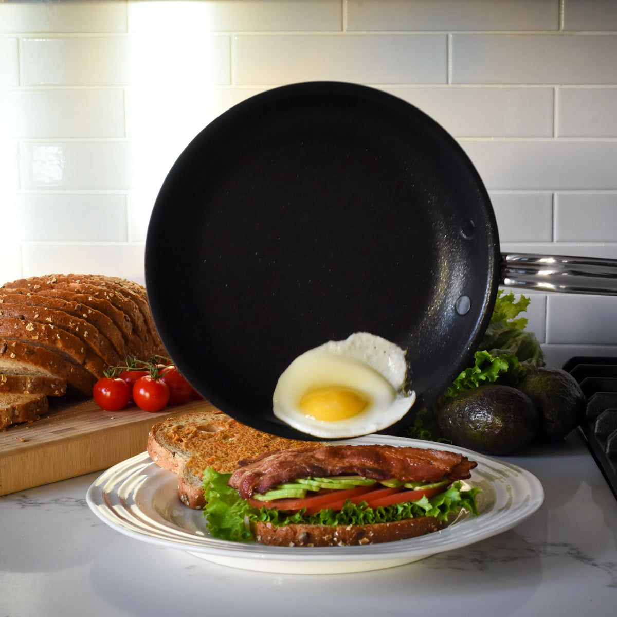 A fried egg is being slid from a ProBond® Forged™ Professional Clad Stainless Steel Epicurean Skillet onto a BLT sandwich on a plate, with sliced bread, tomatoes, avocados, and lettuce visible on the counter in the background.