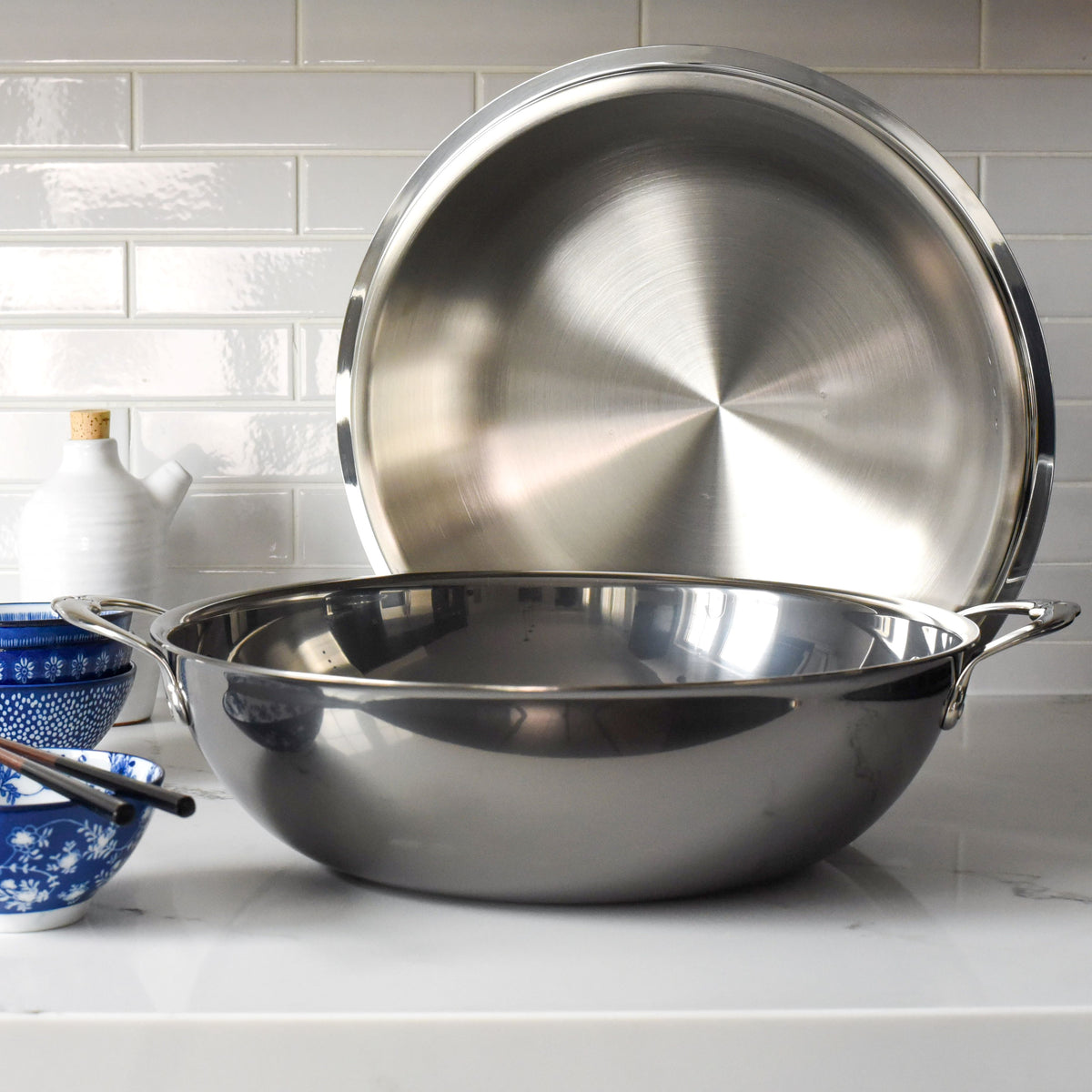 The 14 Stainless Steel Dome Lid by Hestan Provisions sits open on a white kitchen counter against white subway tiles. Nearby, a small blue bowl and sleek white oil bottle enhance its timeless elegance.