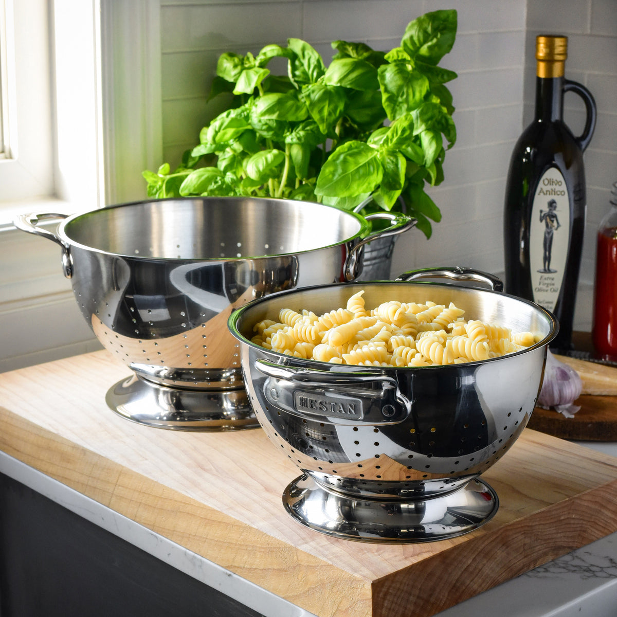 Two Hestan Provisions stainless steel colanders with ergonomic handles rest on a wooden kitchen counter. One is empty, and the other holds cooked pasta. A bottle of olive oil, garlic, and fresh basil are in the background. These dishwasher-safe colanders ensure easy cleaning.