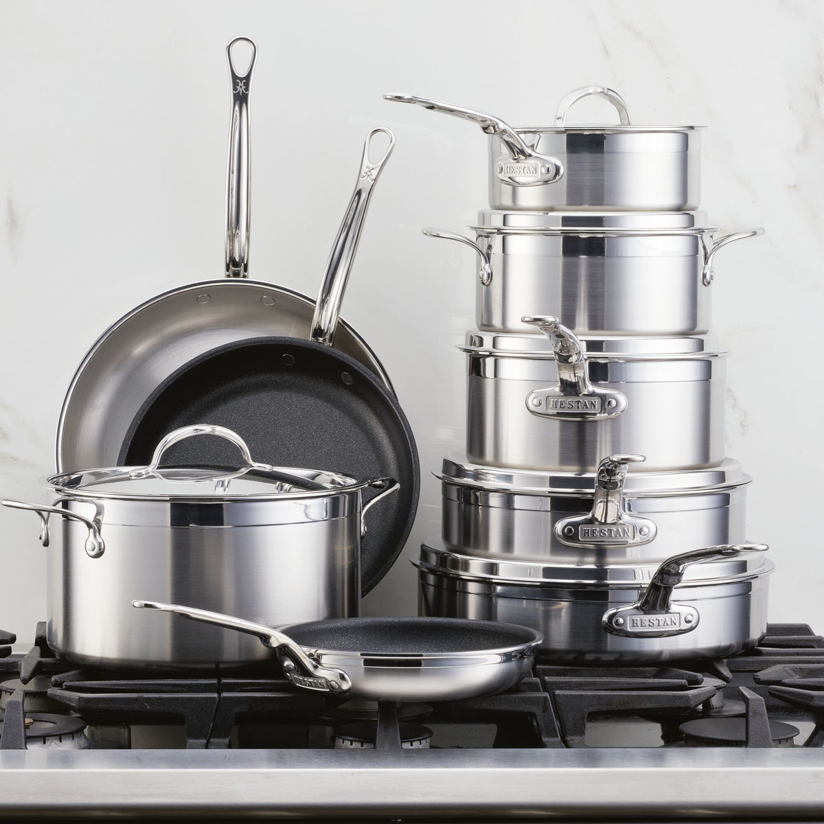 The ProBond® Forged™ Professional Clad Stainless Steel Epicurean Set, 15-piece, with polished surfaces and sturdy handles, is shown on a stovetop with some pots, pans, and lids stacked together.