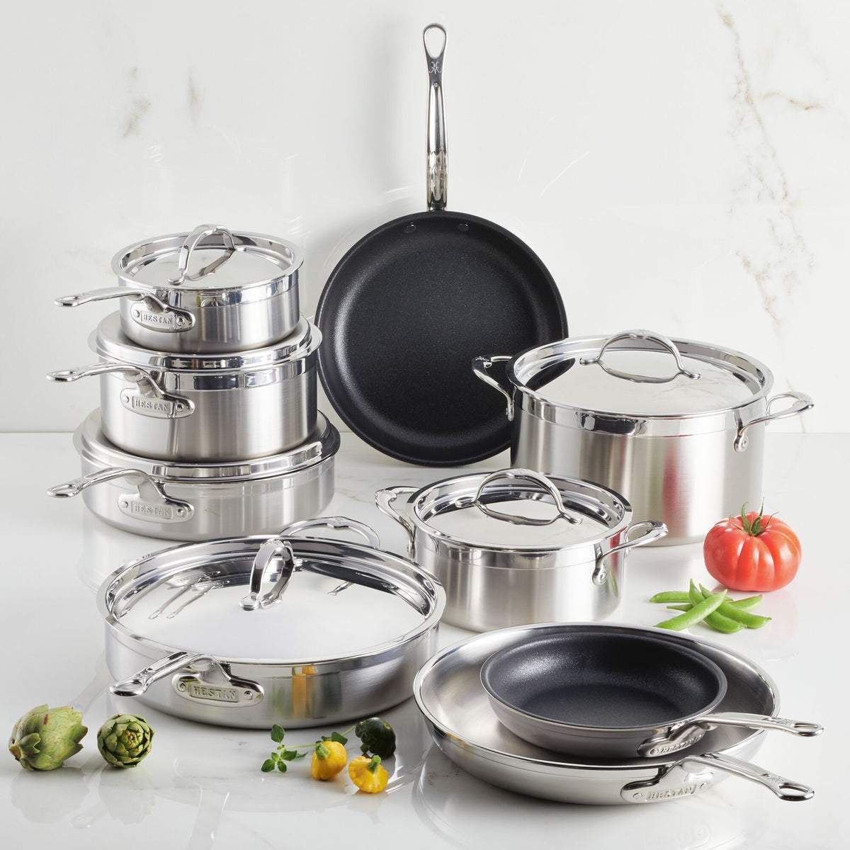A ProBond® Forged™ Professional Clad Stainless Steel Epicurean Set, 15-piece, featuring pots, pans, and lids, is displayed on a white surface surrounded by fresh vegetables like tomatoes, peas, and artichokes.