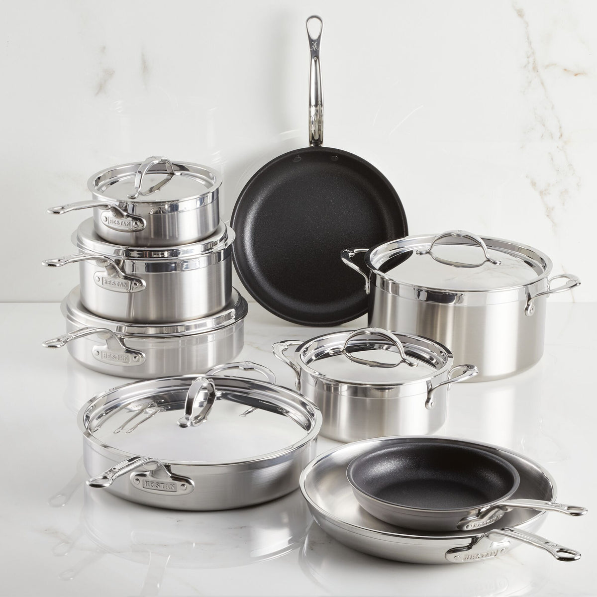 A ProBond® Forged™ Professional Clad Stainless Steel Epicurean Set, 15-piece, including lidded Tri-Ply Body pots, saucepans, and TITUM® Nonstick Skillets, is arranged on a white surface with a white marble wall in the background.